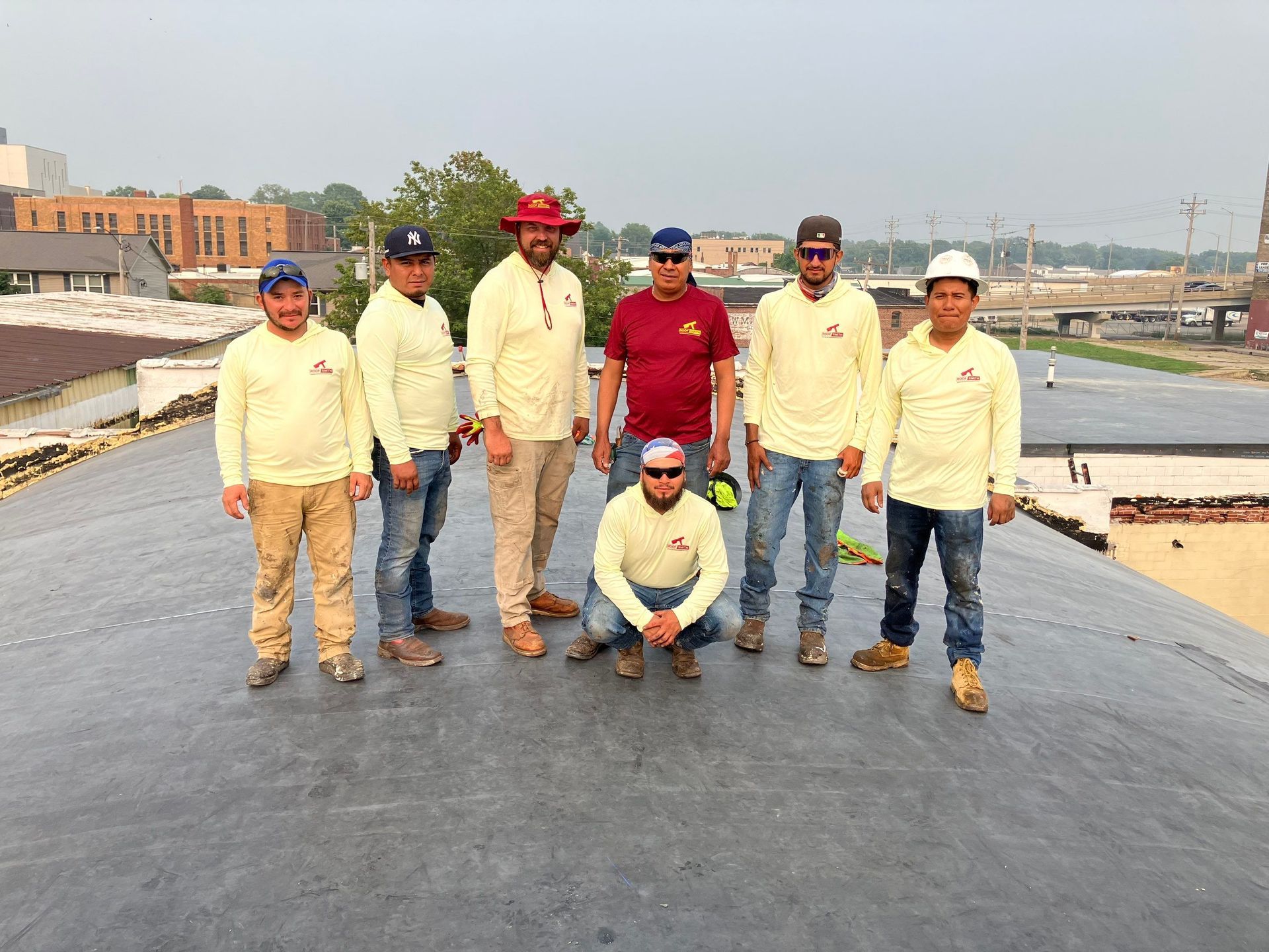 Seven workers posing on a road construction site, with a dark asphalt surface and equipment in the background.