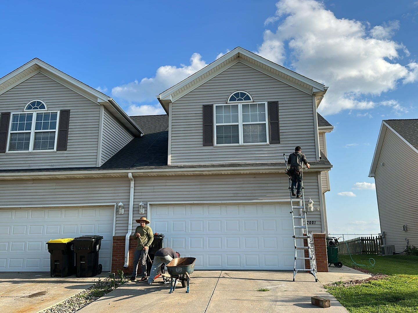 Two workers on ladders painting a gray two-story house with white garage doors