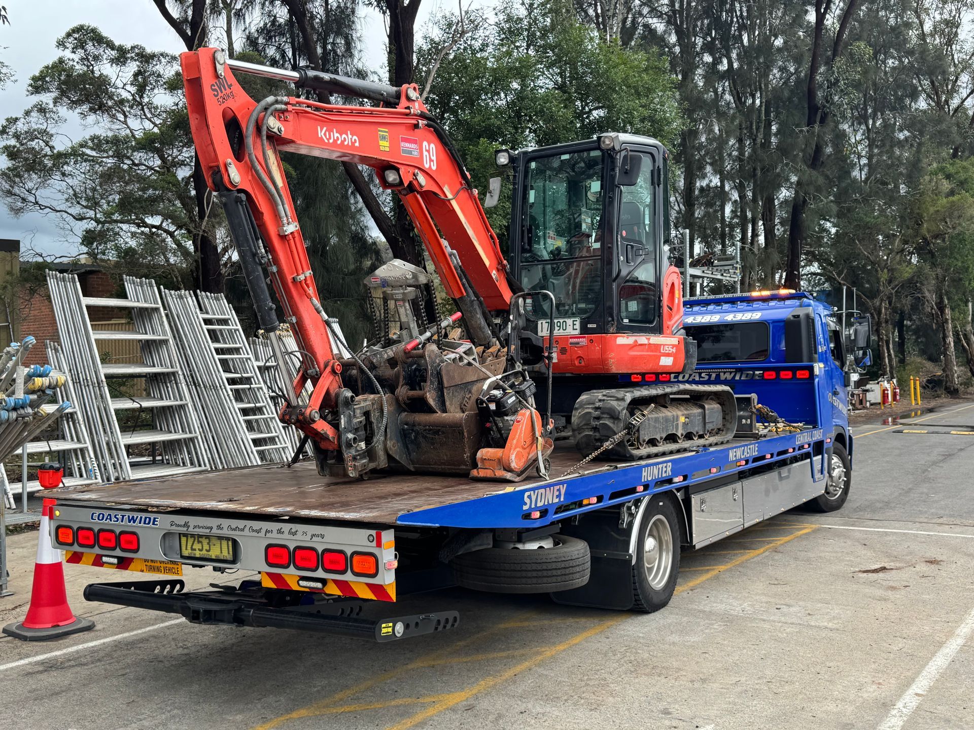 Mini excavator on a blue flatbed truck. Red excavator arm. Street scene with ladders. — Coastwide Towing & Transport In Berkeley Vale, NSW