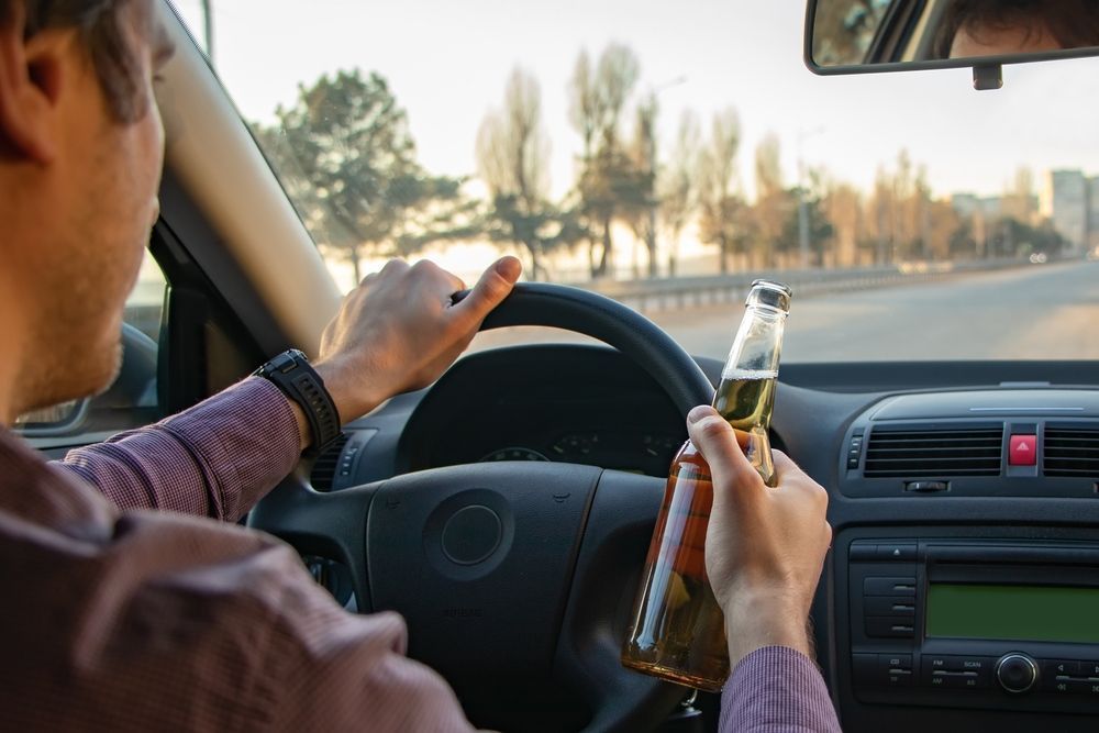 A man is driving a car while holding a bottle of beer.