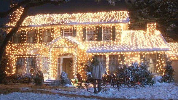 House covered in glowing Christmas lights; snow-covered lawn; person walking toward the house.