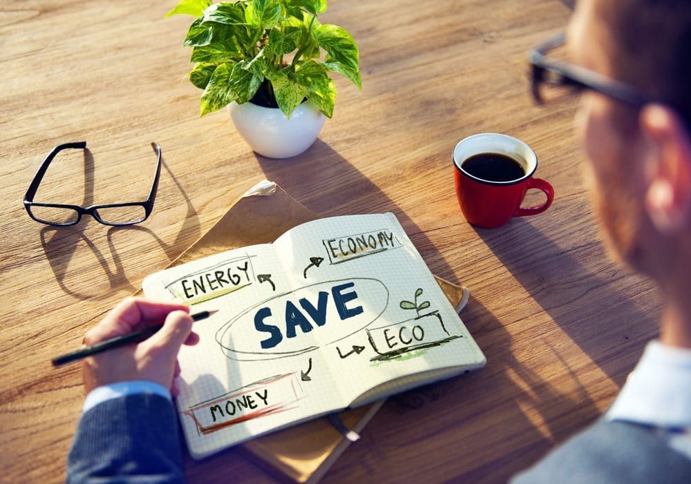 Person sketching “SAVE” concept with “Energy,” “Economy,” “Eco,” and “Money” on notepad; coffee and plant nearby.