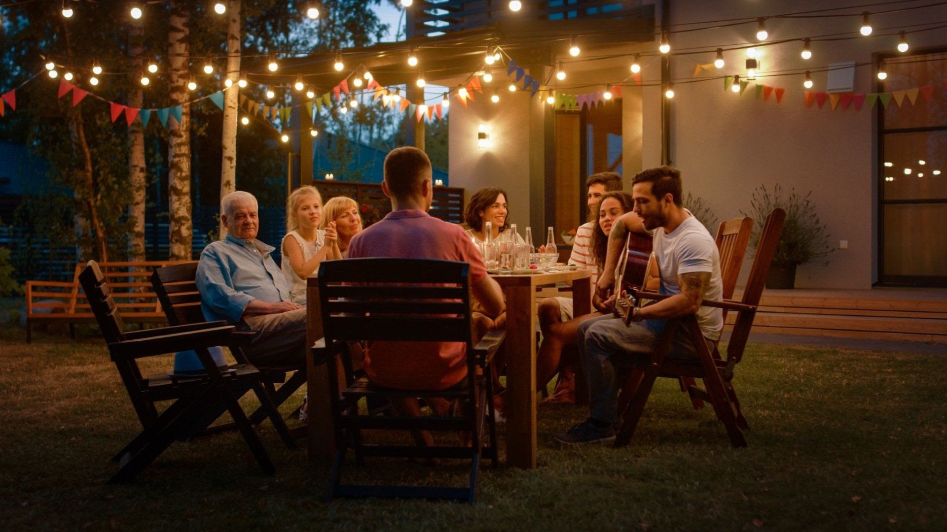 People gather around a table outdoors at night, illuminated by string lights.