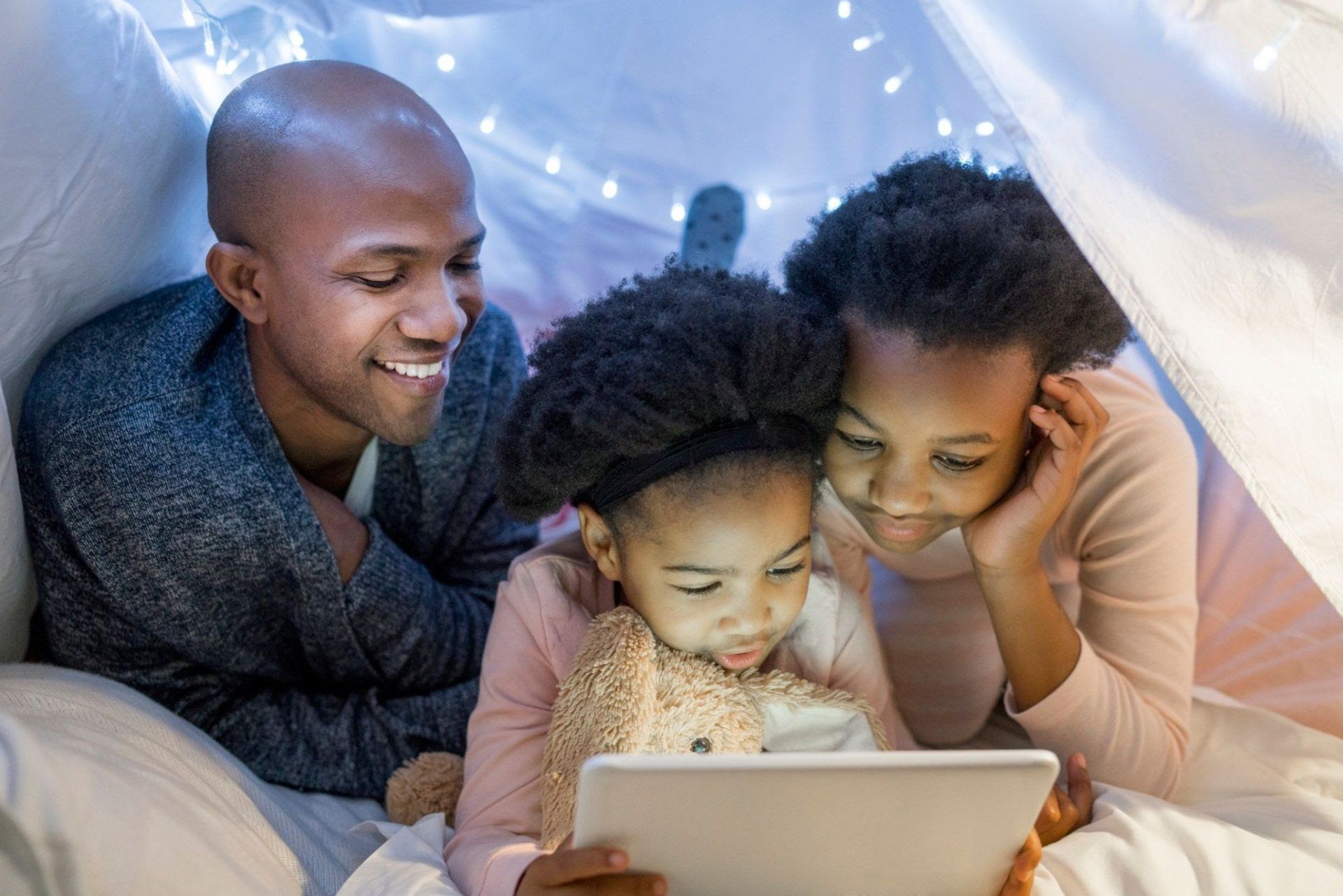 Family looks at a tablet inside a blanket fort, lit with fairy lights.