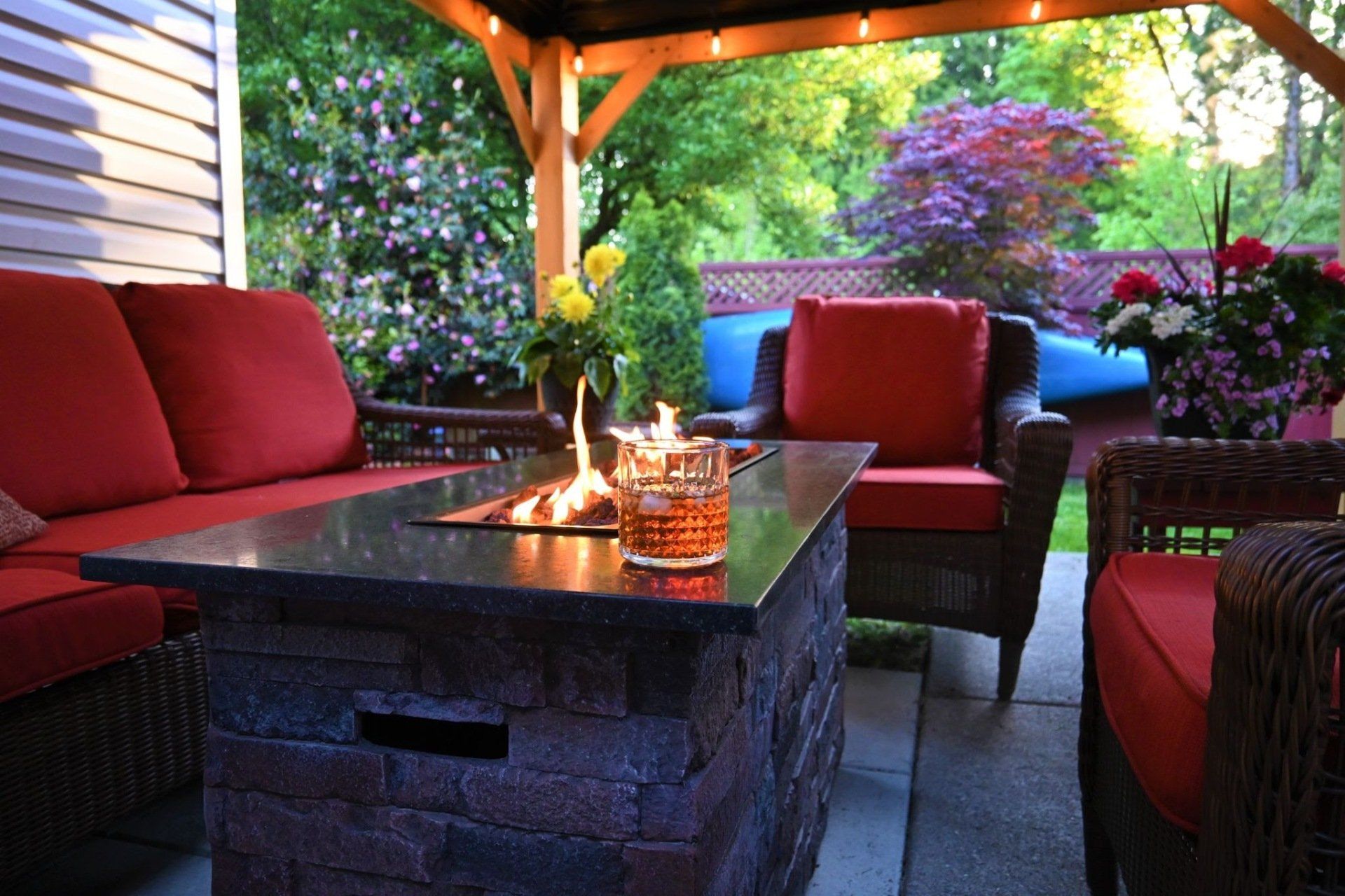 Outdoor patio with fire pit, red cushioned furniture, under a wooden gazebo with lush green backdrop.