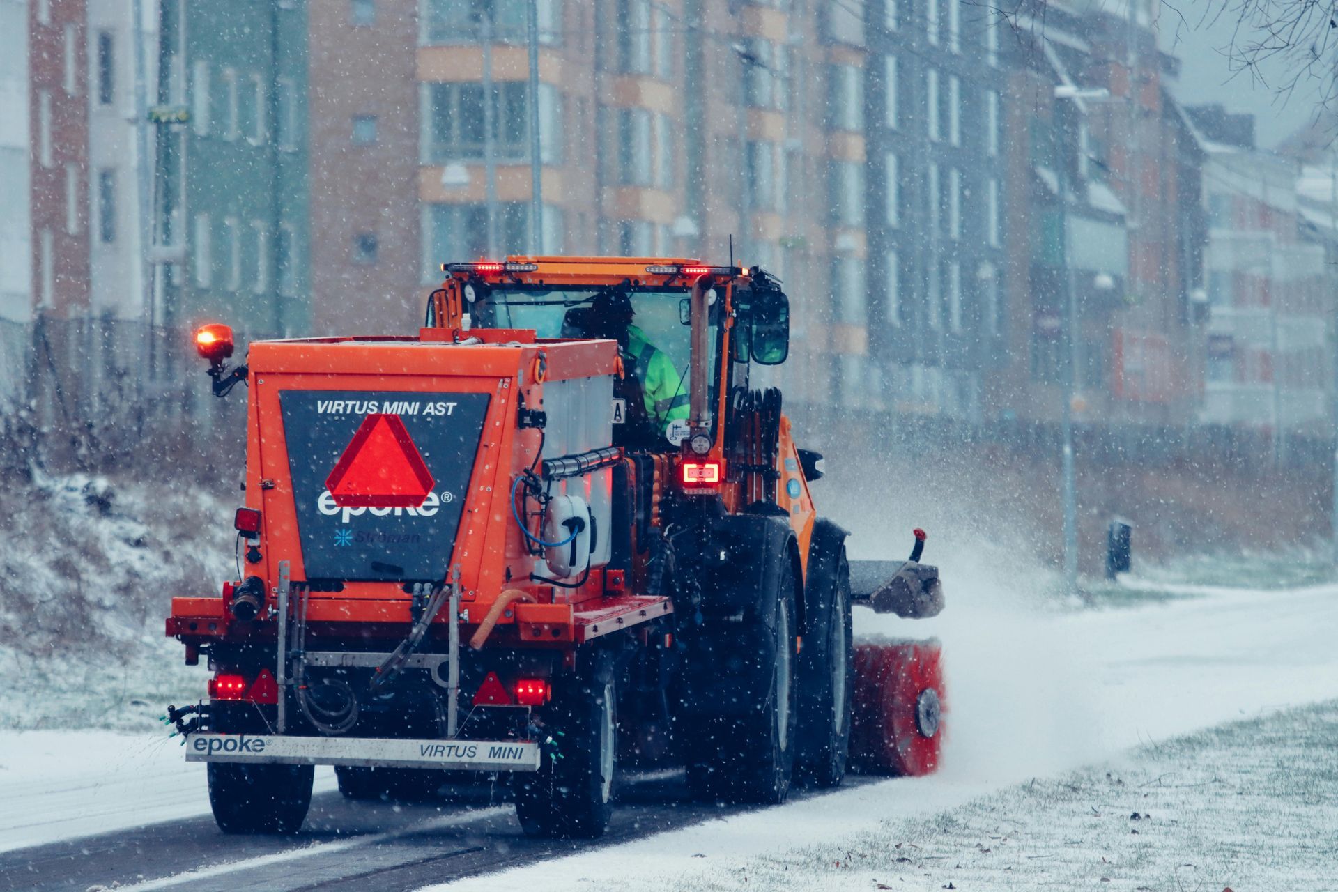 Orange snowplow clearing a snowy road; buildings in the background.