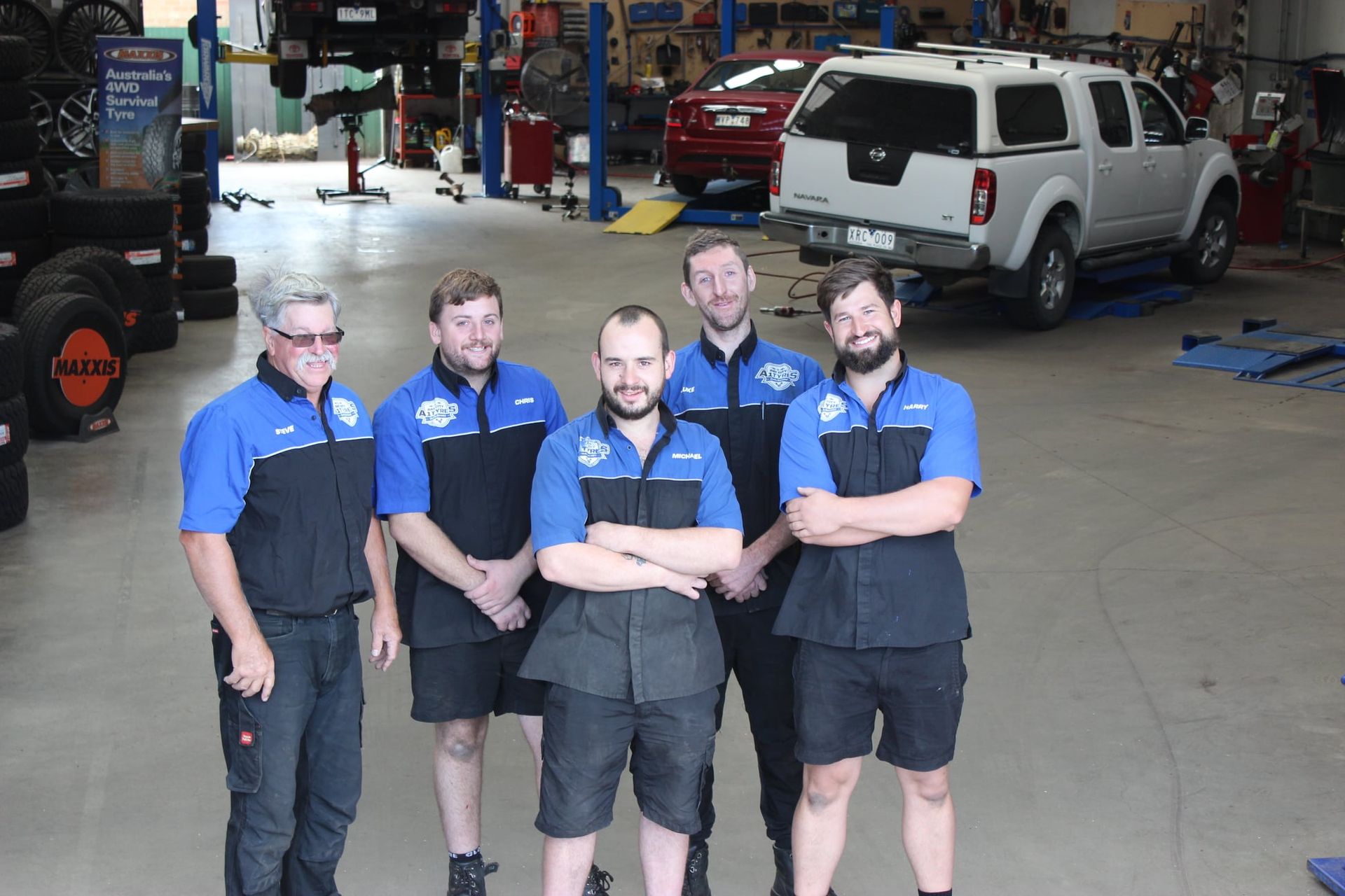 Five mechanics in blue shirts stand in an auto repair shop with a white SUV on a lift behind them.