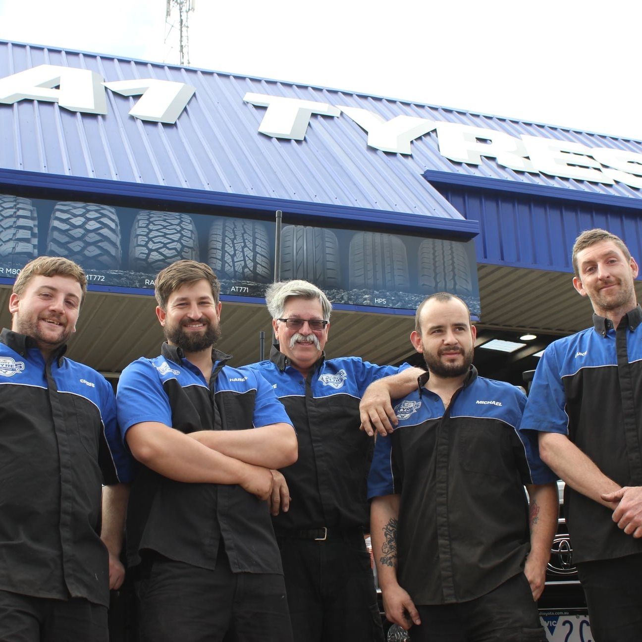 Five workers in blue-and-black uniforms standing outside a building with a blue roof, arms crossed.