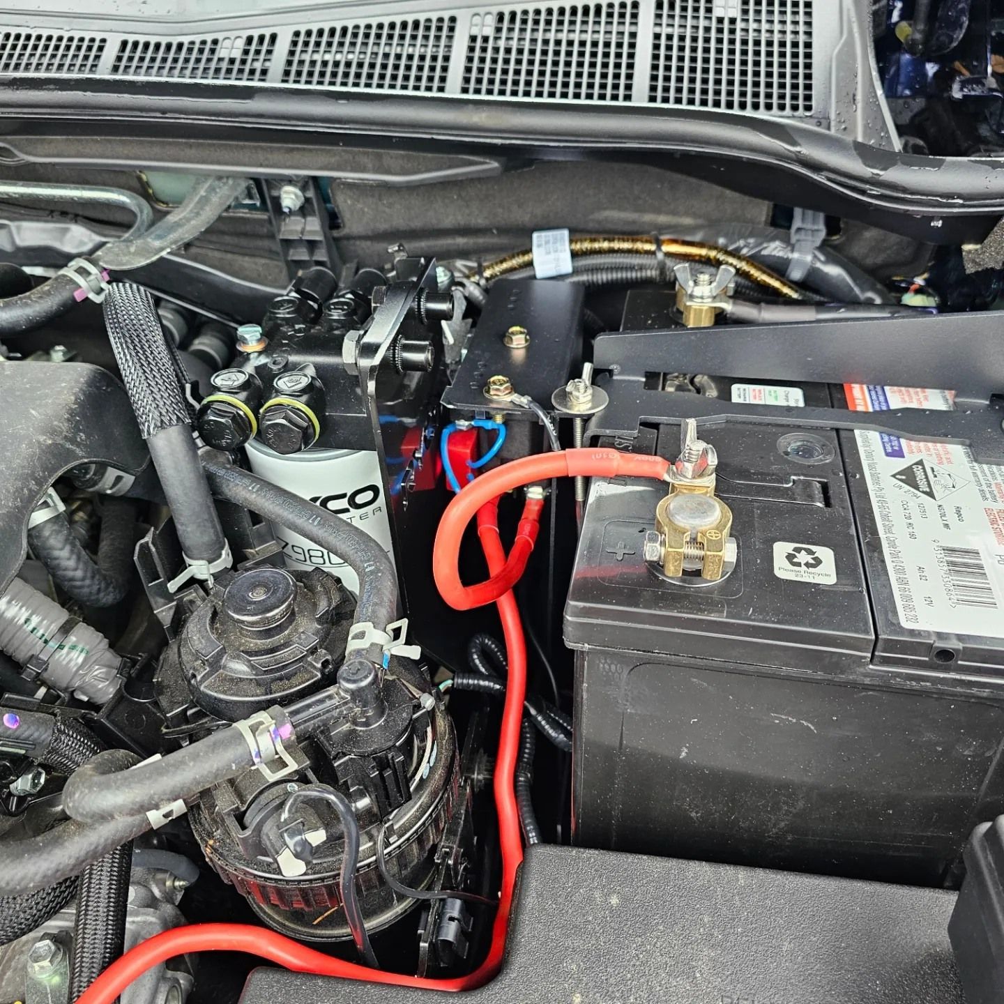 Car engine bay with black battery and red cables, showing electrical wiring and components.