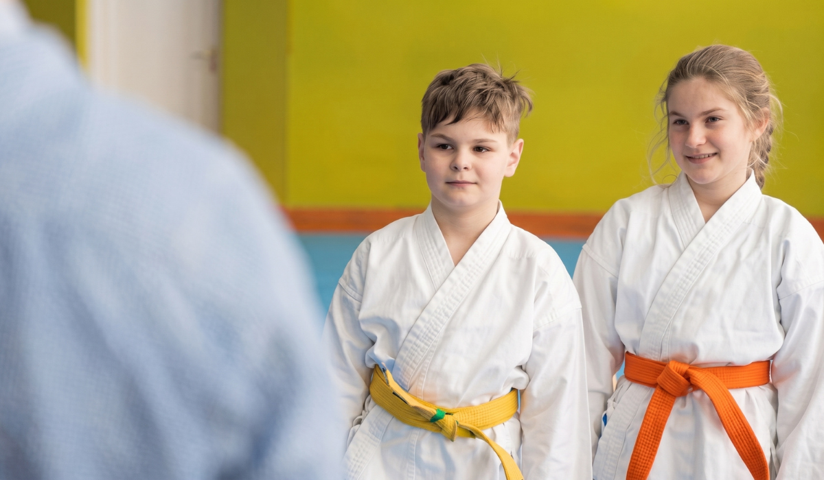Two children in white martial arts uniforms with colored belts standing in a dojo