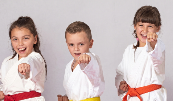 Three children in white martial arts uniforms with colored belts, posing and pointing toward the camera.