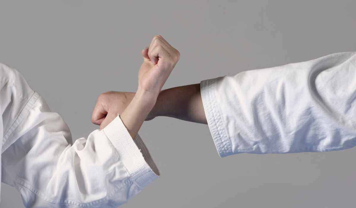 Two people in white martial arts uniforms crossing forearms against a plain gray background