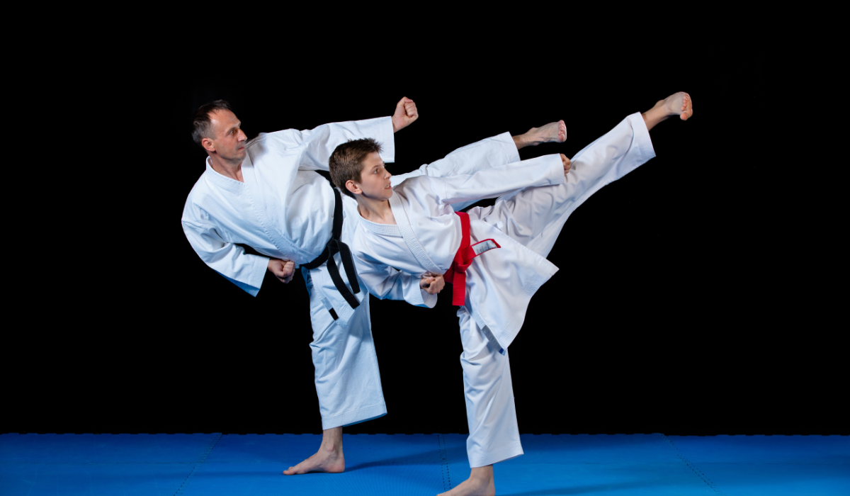 Two martial artists in white uniforms sparring, one delivering a high side kick on a blue mat against a black background