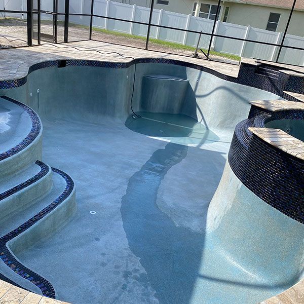 Empty blue-tiled swimming pool with curved steps and a raised spa area, surrounded by a glass fence.