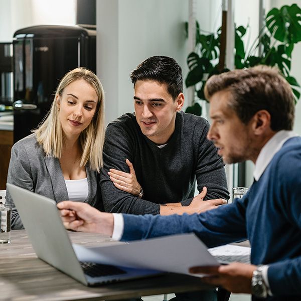 Three coworkers review documents together at a laptop in a modern office.