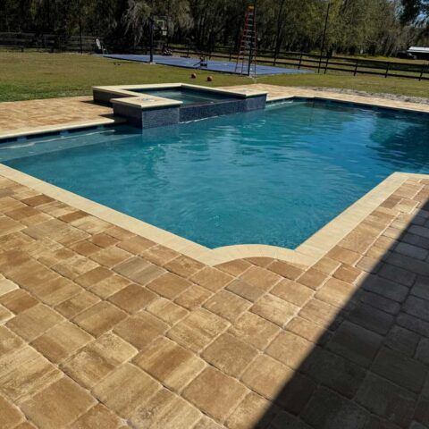 Backyard swimming pool with tan stone deck and raised water feature, surrounded by trees and fence.