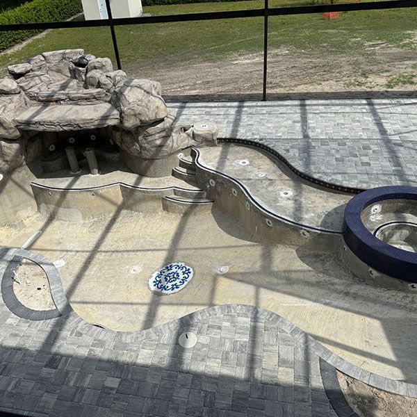 Empty swimming pool with stone feature, blue drain cover, and tiled deck behind a black fence