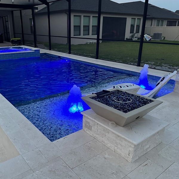 Backyard pool with blue-lit fountains and a square fire pit on a stone patio