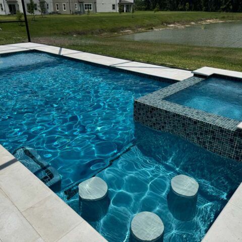 Modern backyard pool with blue water, submerged stools, and an elevated spa overlooking a grassy pond.
