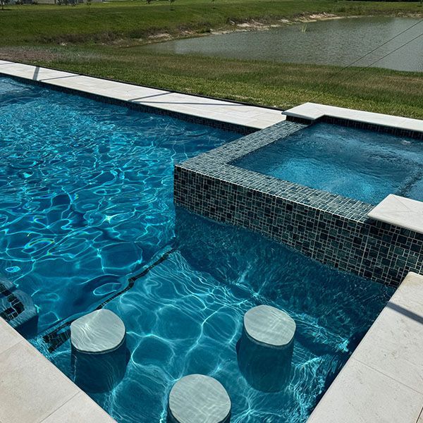 Modern pool with turquoise water, square spa, and three round stools beside a grassy lakeside view