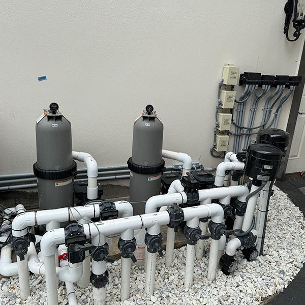 Outdoor pool filtration equipment with white PVC pipes, gray tanks, and valves on gravel against a wall