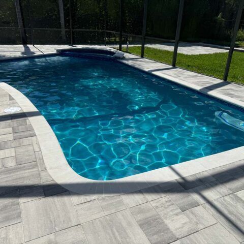 Rectangular backyard swimming pool with clear blue water and light stone deck, surrounded by a safety fence