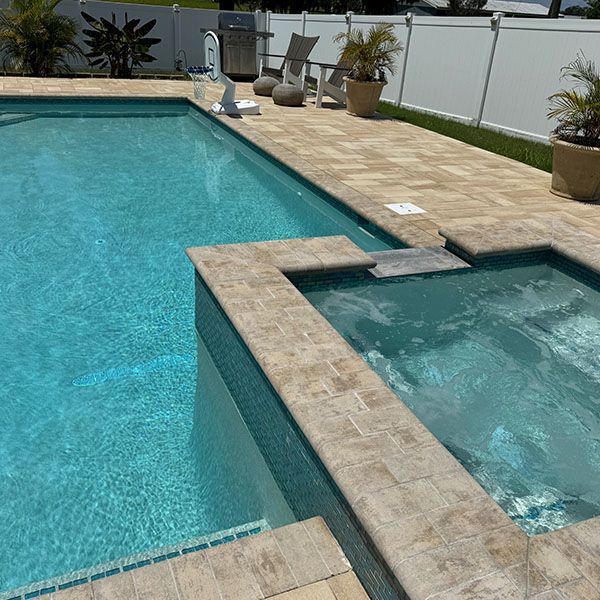 Backyard swimming pool with attached hot tub and tan stone deck beside white fence