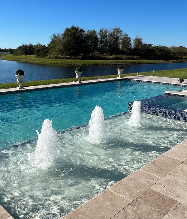 Outdoor pool with bubbling fountains beside a calm lake under a clear blue sky