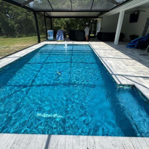 Long rectangular backyard pool with bright blue water under a screened patio and covered seating area