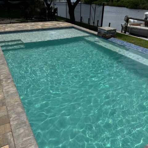 Backyard swimming pool with clear turquoise water beside a dock on a lake.