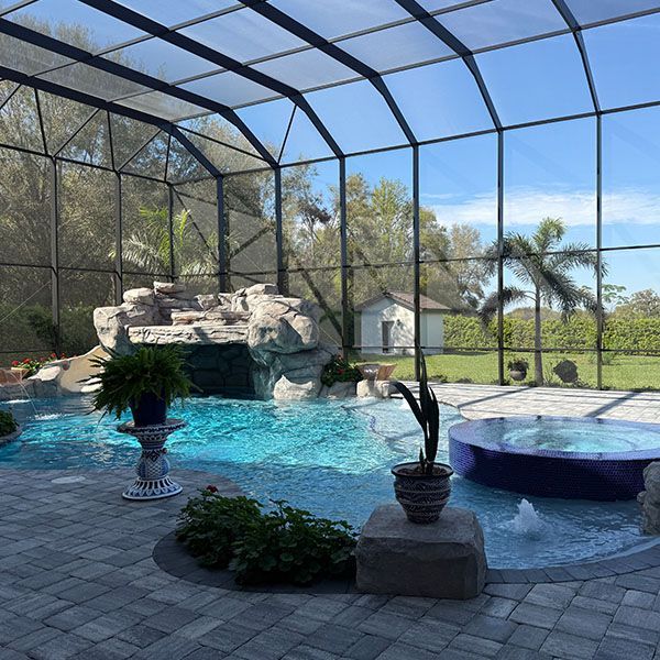 Screened-in backyard pool with rock waterfall, spa, and potted plants under a blue sky