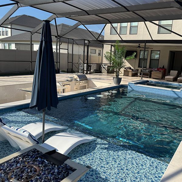 Backyard pool under a covered patio with lounge chairs, umbrella, and a fire pit beside a house