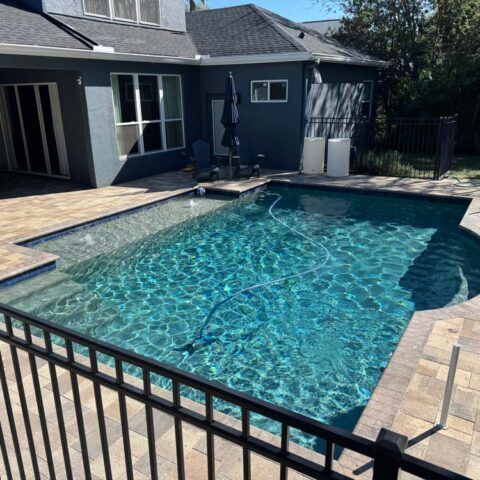 Backyard swimming pool with clear blue water, stone deck, and patio beside a house.
