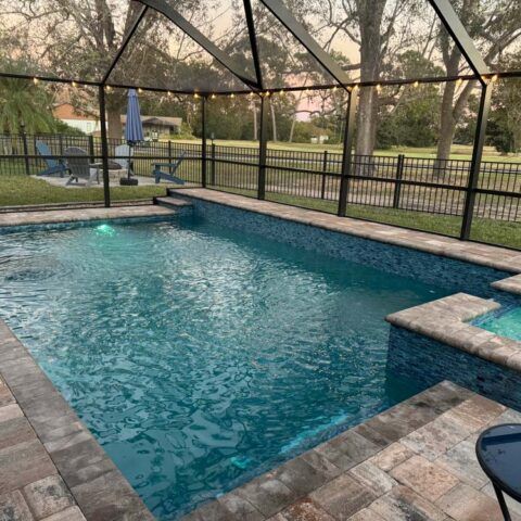 Backyard pool with stone deck, black fence, and screened pergola at dusk