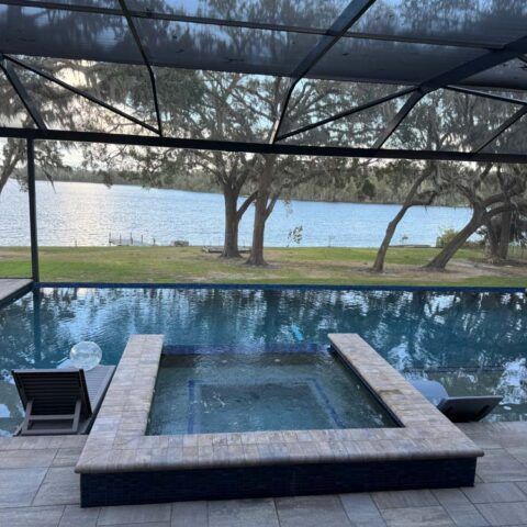 Infinity pool and hot tub overlooking a calm lake at sunset, framed by patio chairs and trees.