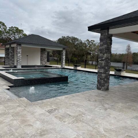 Modern backyard pool with a raised spa, stone pavers, and covered pavilion under cloudy skies