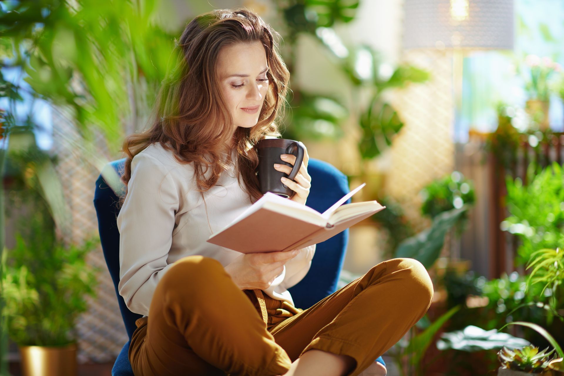 Une femme lit un livre en sirotant une boisson dans une tasse, entourée de plantes dans un espace bien éclairé.