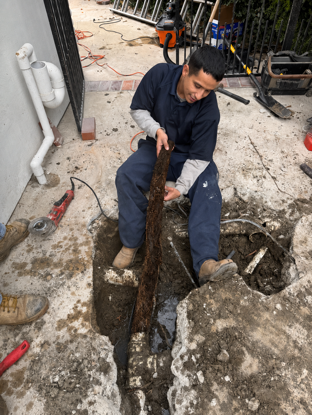 Worker in dark coveralls digging a trench with a long tool beside exposed pipes and broken concrete.