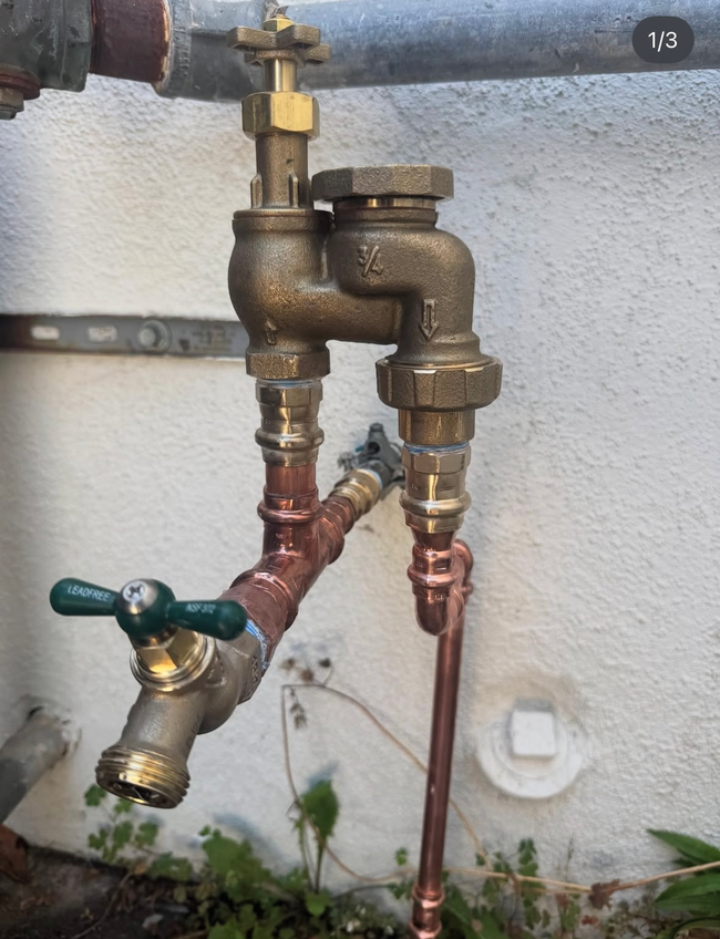 Outdoor plumbing with two connected brass water valves and pipes against a white wall.