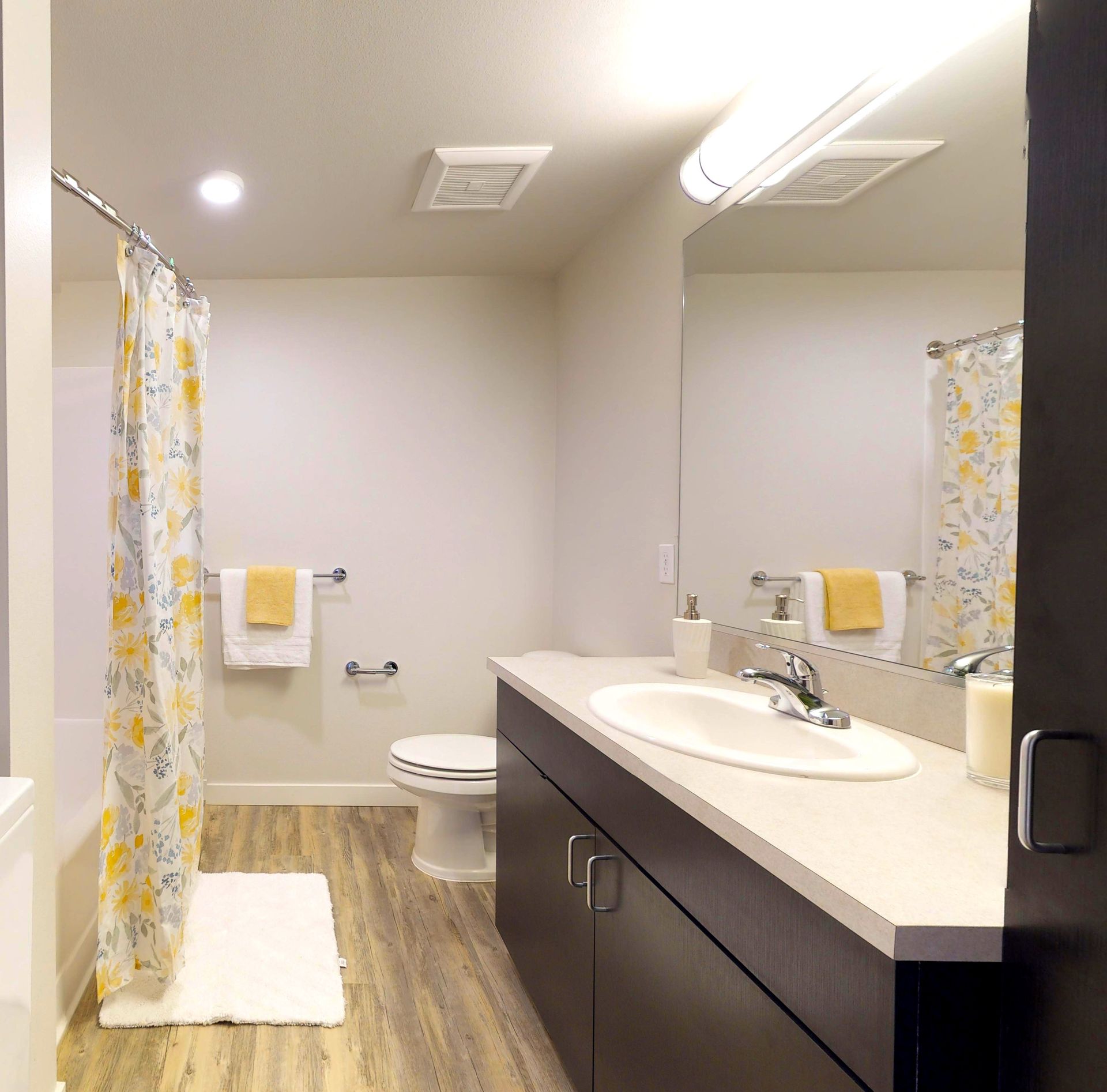 Bathroom with white walls, a gray-toned wood floor, and a dark brown vanity with a white countertop.