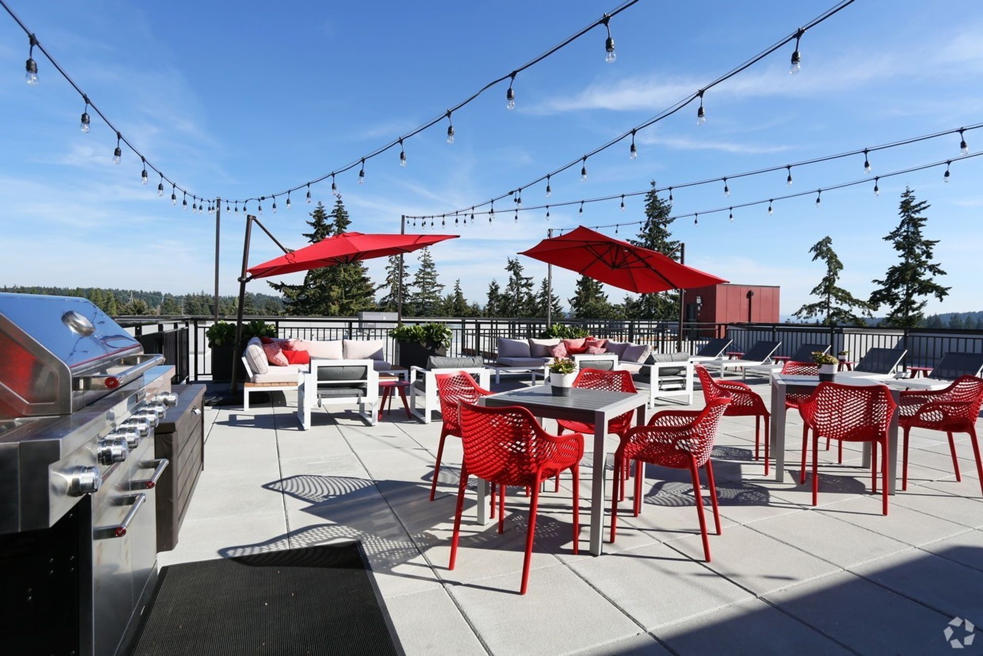 Rooftop patio with grill, red umbrellas, tables, chairs, and string lights against a blue sky.
