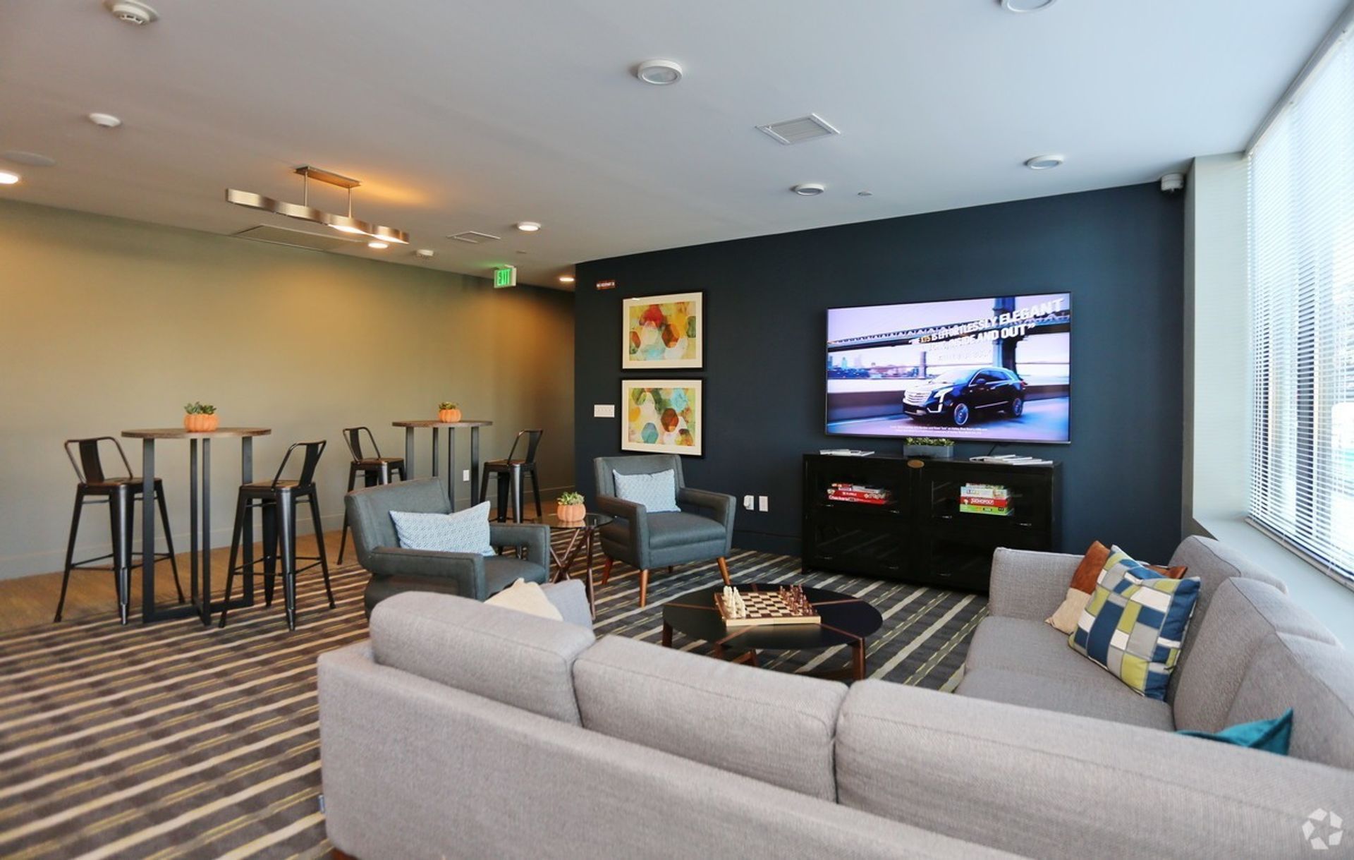 Modern lounge area: gray sofa, armchairs, high-top tables, large TV on dark blue wall, striped rug.