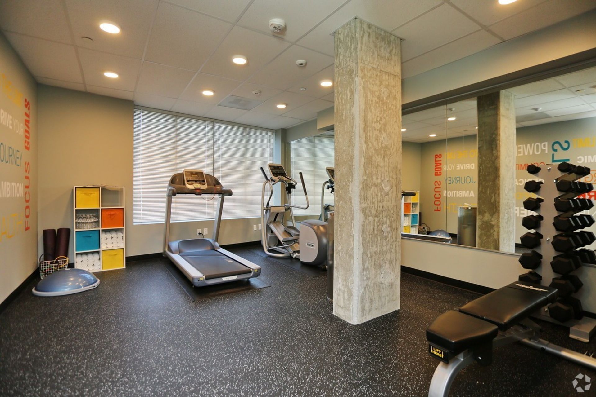 Gym with treadmill, elliptical, free weights, and balance ball. Black rubber flooring, column, and mirror.