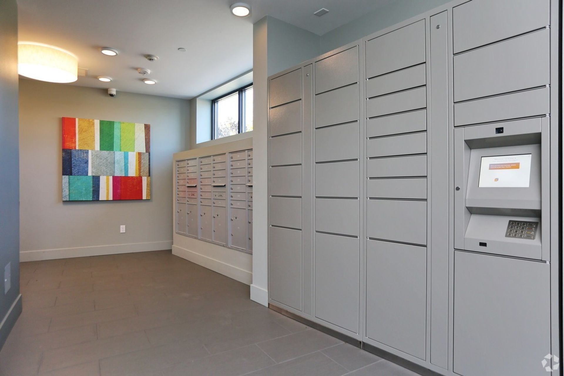 Lobby with mailboxes and package lockers. A colorful painting hangs on a wall; a touchscreen kiosk is on the right.