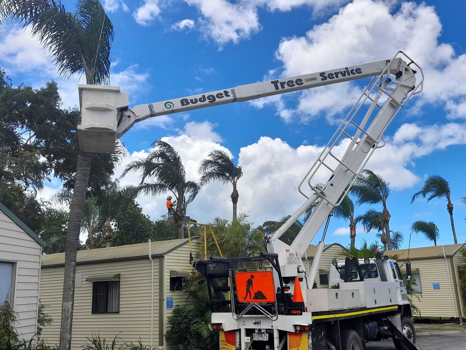 Workers in orange safety gear in a cherry picker cutting a tree branch outdoors