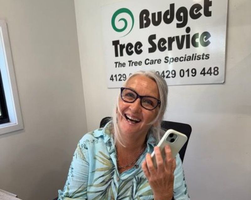 Smiling woman holding a phone in an office beside a Budget Tree Service sign