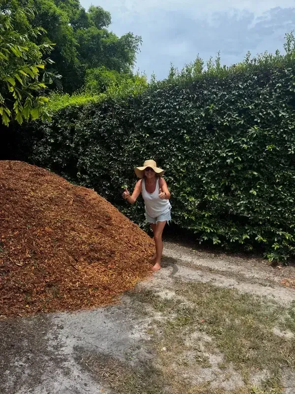 Woman in a sun hat standing beside a large pile of mulch on a gravel path near tall hedges and trees