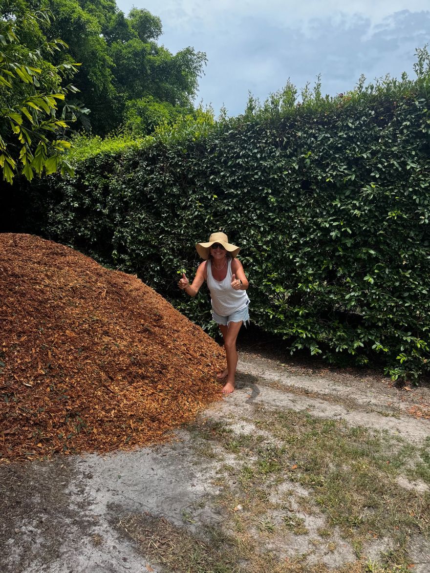 Person standing beside a large pile of leaves on a gravel path, wearing a sun hat in a leafy garden
