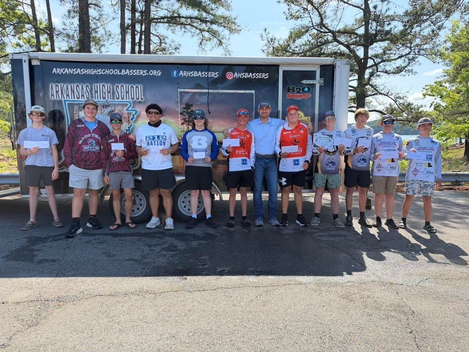 Group of cyclists posing in front of a support trailer on a sunny roadside.