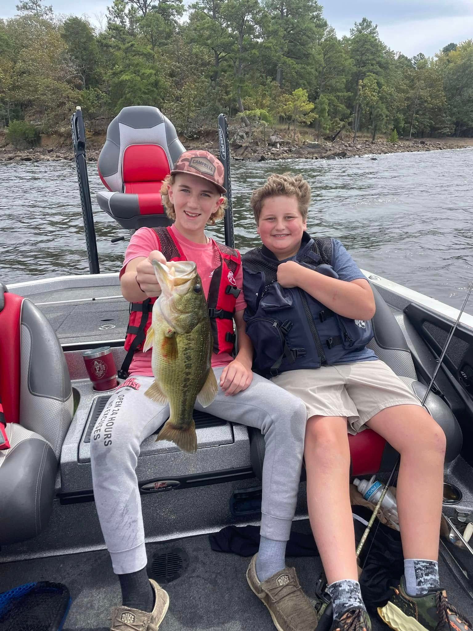 Two people on a boat holding a large fish, with a lake and trees in the background.